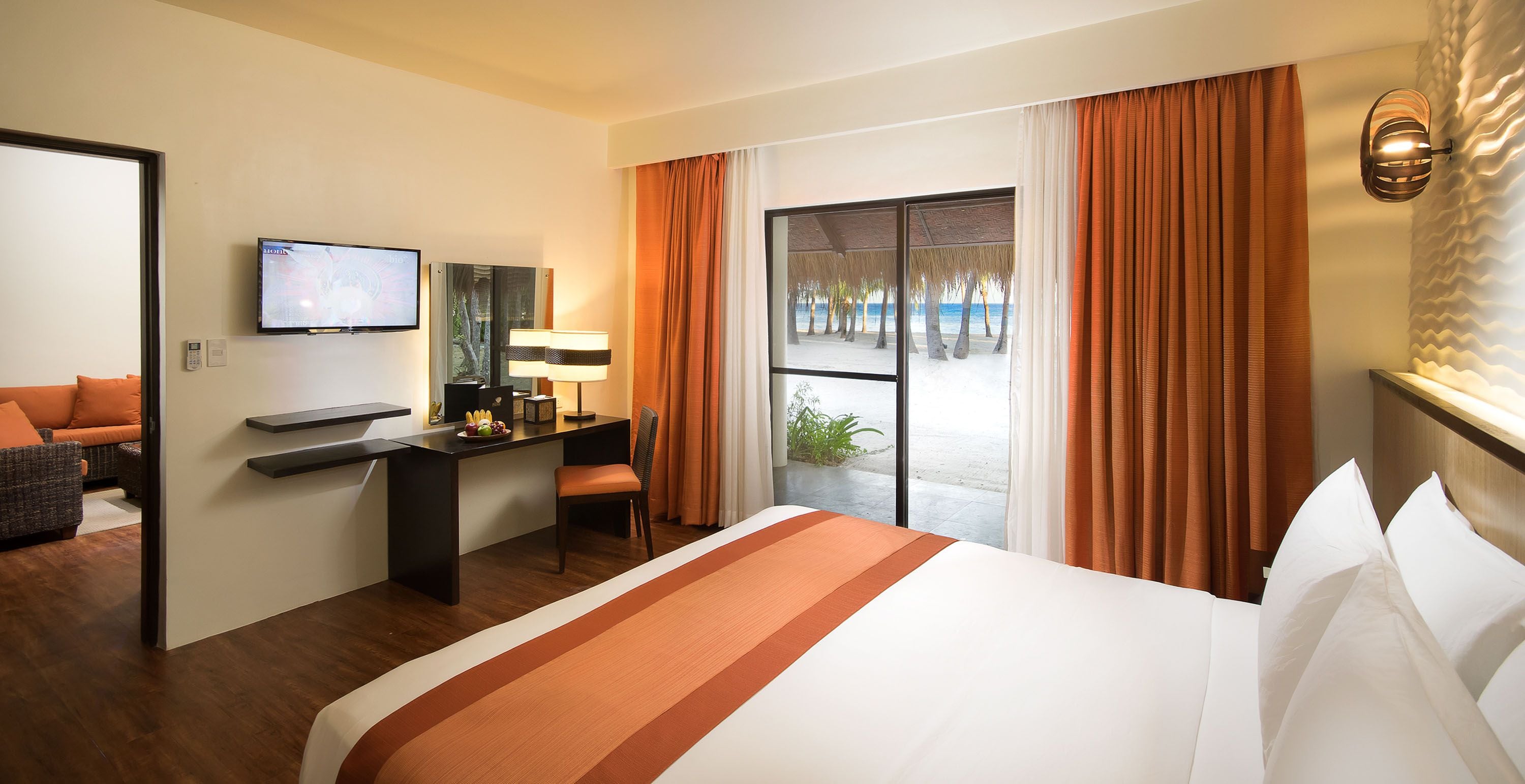 A hotel room with a bed, a desk, a chair, a TV, and a view of the beach.
