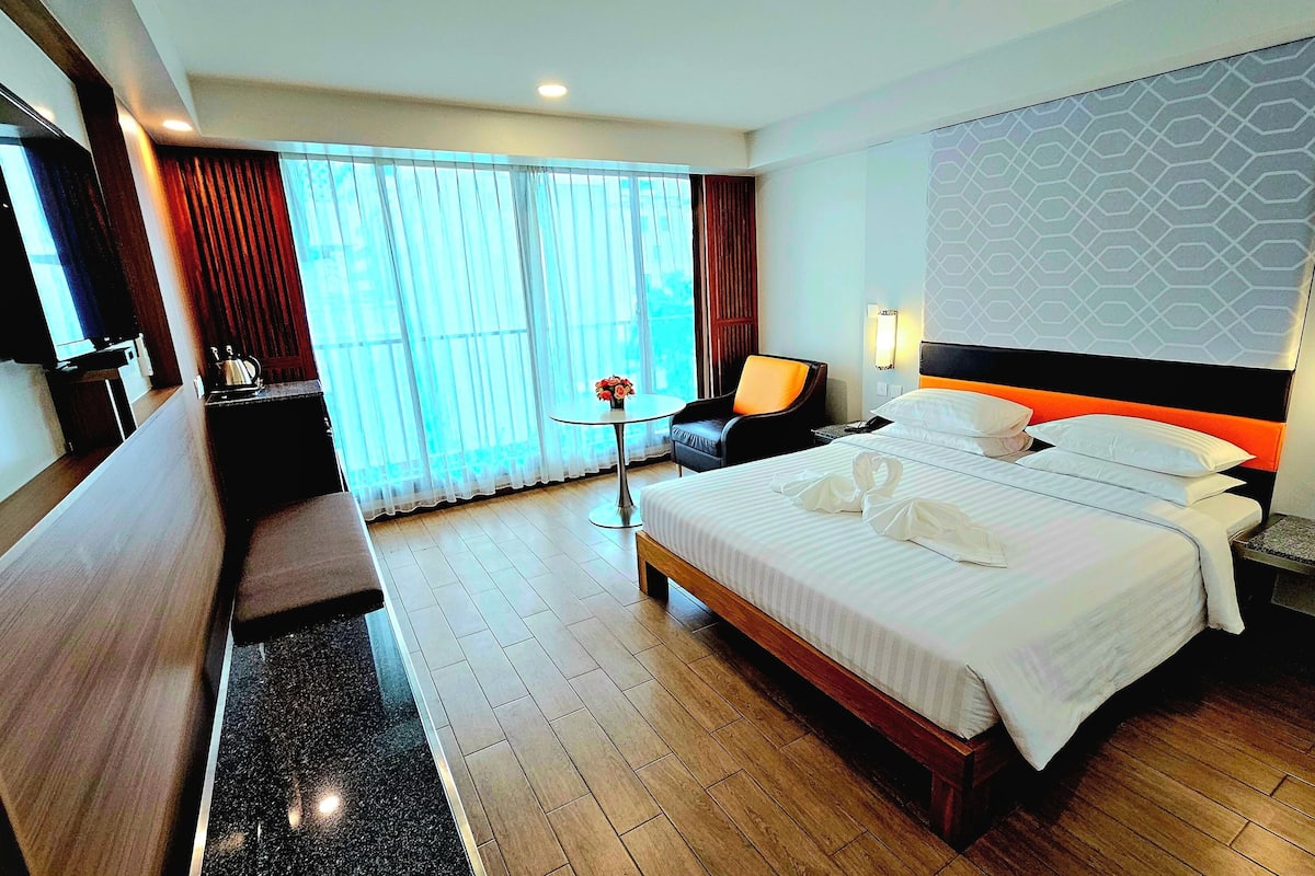 A hotel room with a large bed, a desk, a chair, a small table with flowers, and a view of the outdoors through the window.