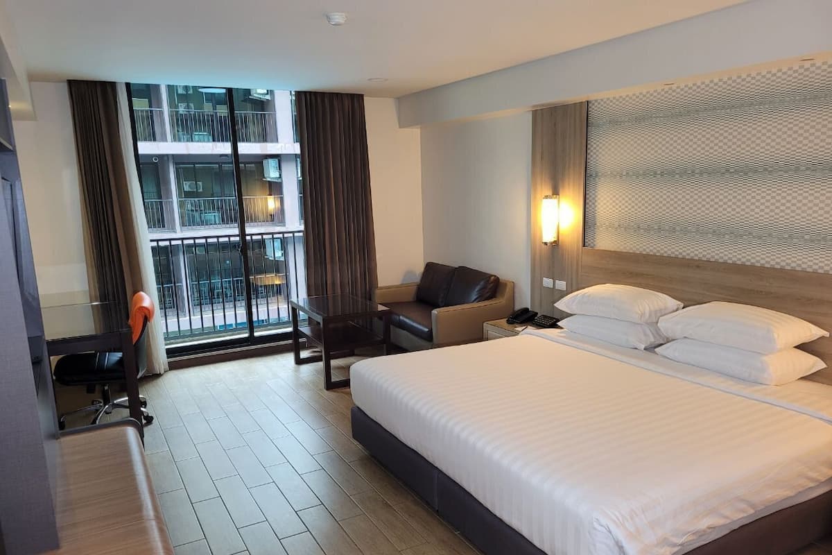 A hotel room with a large bed, a desk, a chair, a sofa, and a view of a building through a large window.