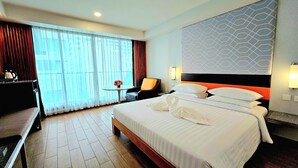 A hotel room with a large bed, a desk with a chair, a small table with flowers, and a window with curtains. - Dynasty Grande Hotel (Bangkok)