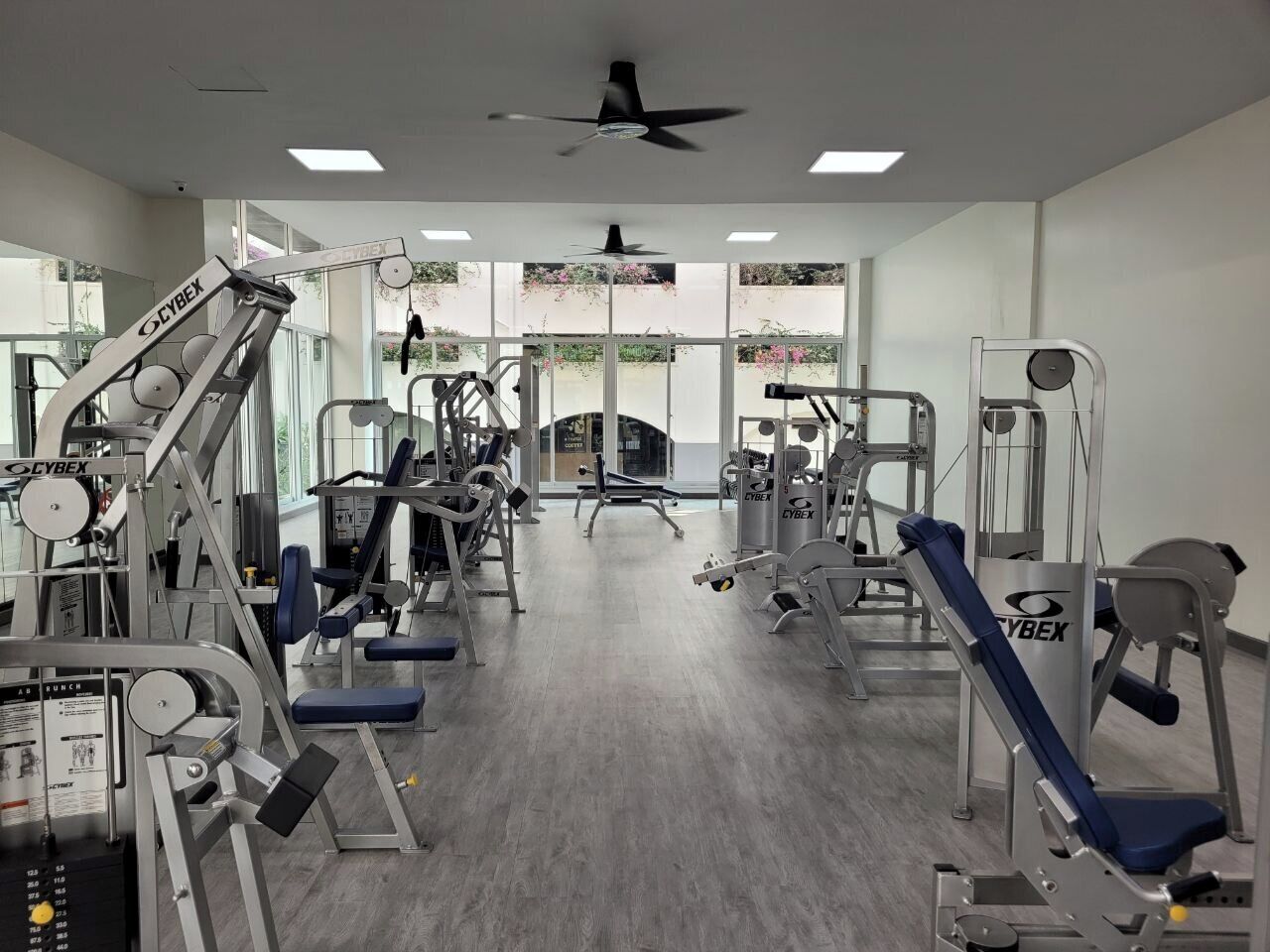 Une salle de sport équipée de divers appareils d’exercice, dotée de grandes fenêtres et d’un ventilateur de plafond.