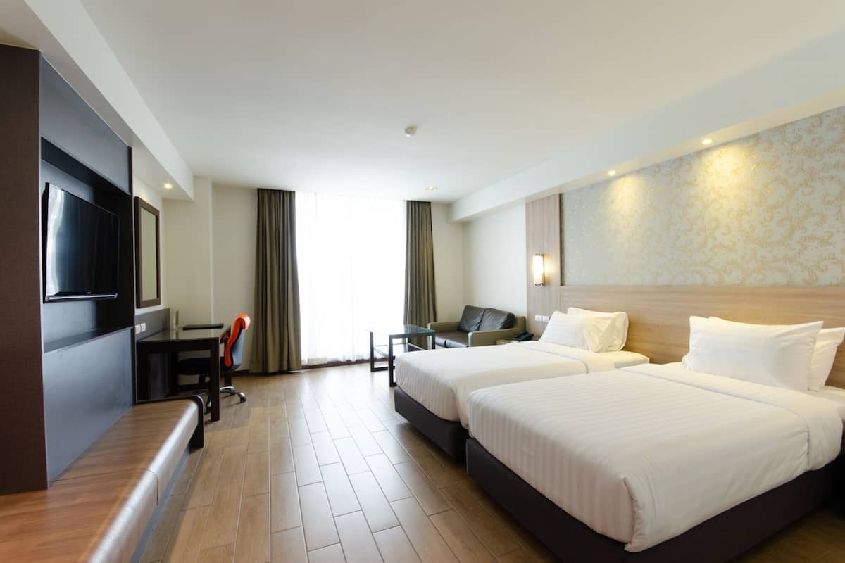 A hotel room with two beds, a desk, a chair, a TV, and a bench.