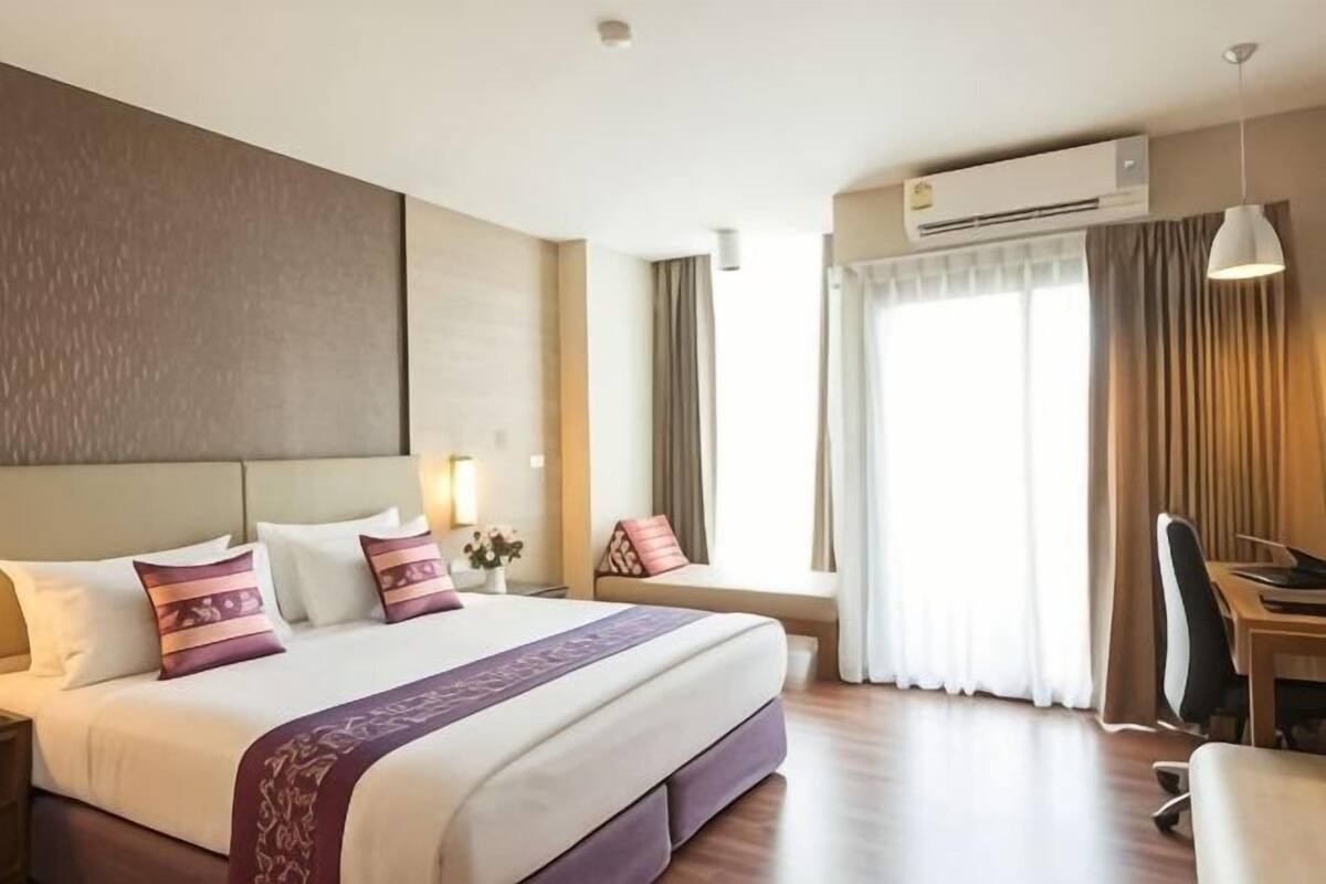 A hotel room with a large bed, a desk, a chair, a window with curtains, and an air conditioning unit.