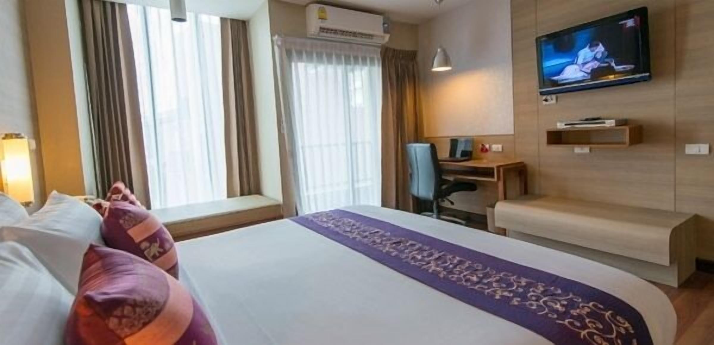 A hotel room with a large bed, a desk, a chair, a TV mounted on the wall, and a window with curtains.