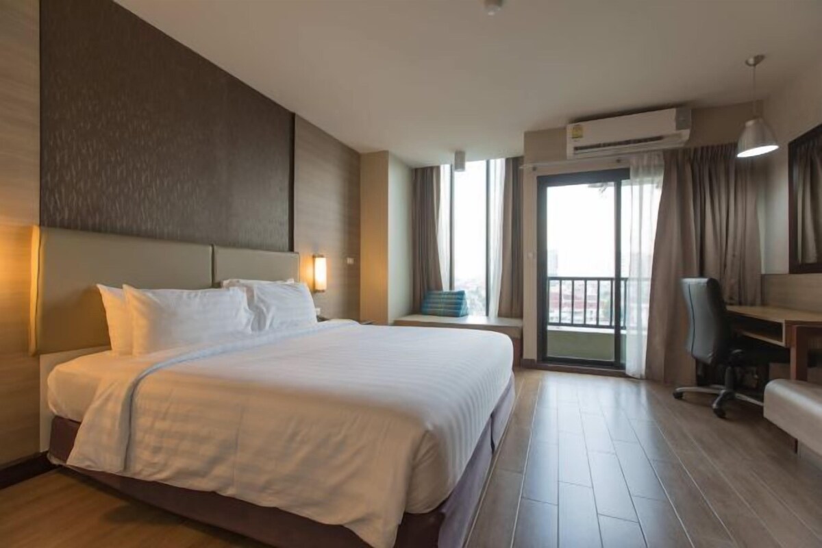 A hotel room with a large bed, a desk, a chair, an air conditioning unit, and a view of the outdoors.