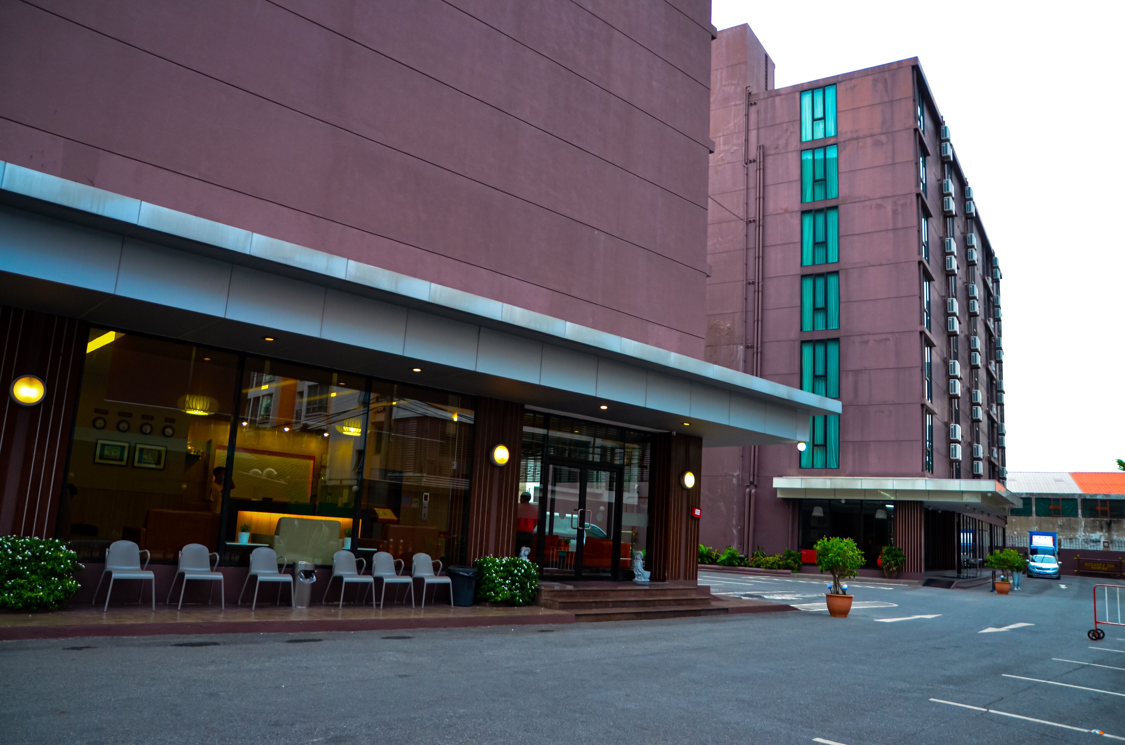 A modern hotel with a red facade, large glass windows, and a covered outdoor seating area with chairs and potted plants.