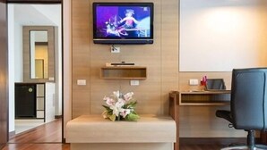 A hotel room with a flat-screen TV mounted on the wall, a small bench with flowers, a desk with a chair, and a wooden cabinet. - Marsi Hotel (Bangkok)