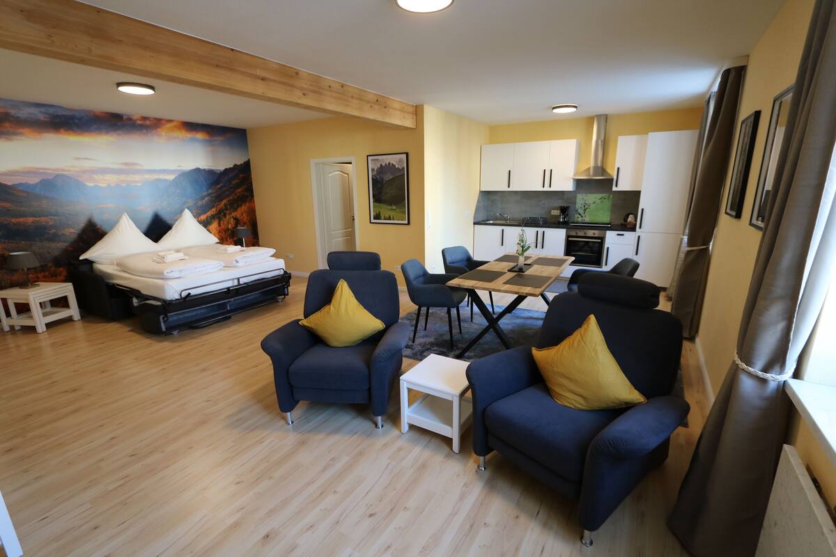 Una habitación de hotel compacta con una cama, dos sillones, una mesita, un rincón de comedor y una cocina.