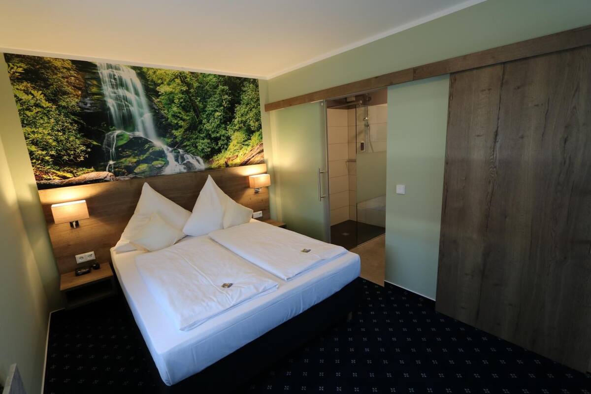 Habitación de hotel con cama, mesita de noche, lámpara de pared, zona de ducha y un espejo grande.