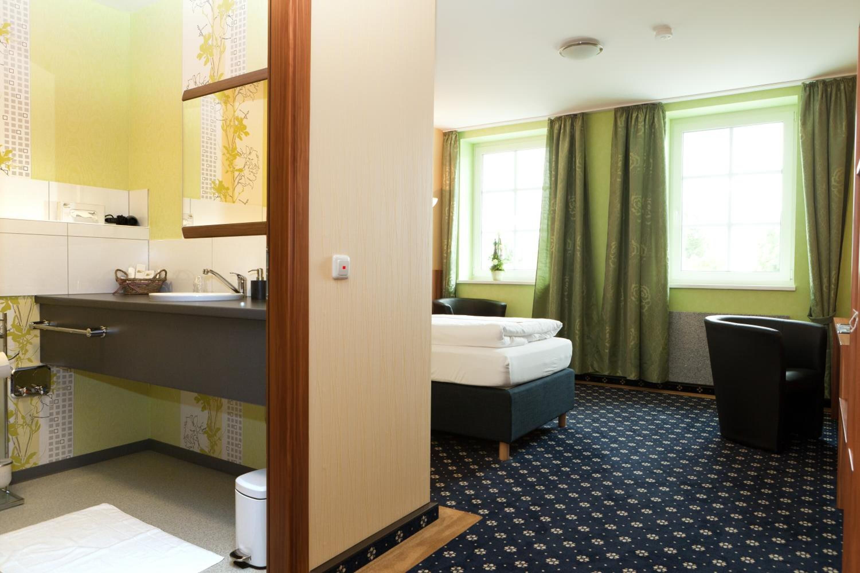 A hotel room with a bed, a desk, a chair, a window with curtains, a sink, and a mirror.