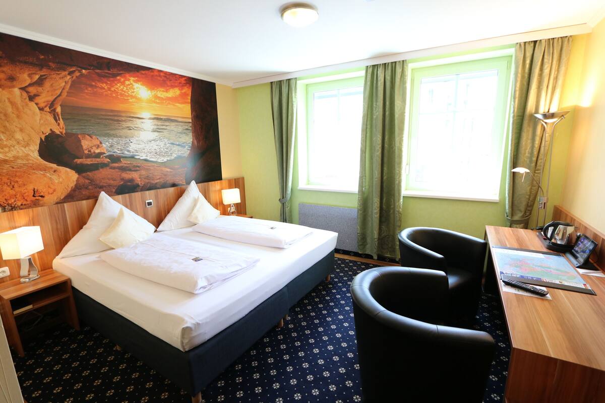 Habitación de hotel con cama, escritorio, silla, lámpara y un cuadro grande con un atardecer sobre el océano.