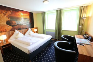 Habitación de hotel con cama, escritorio, silla, lámpara y un cuadro grande con un atardecer sobre el océano.