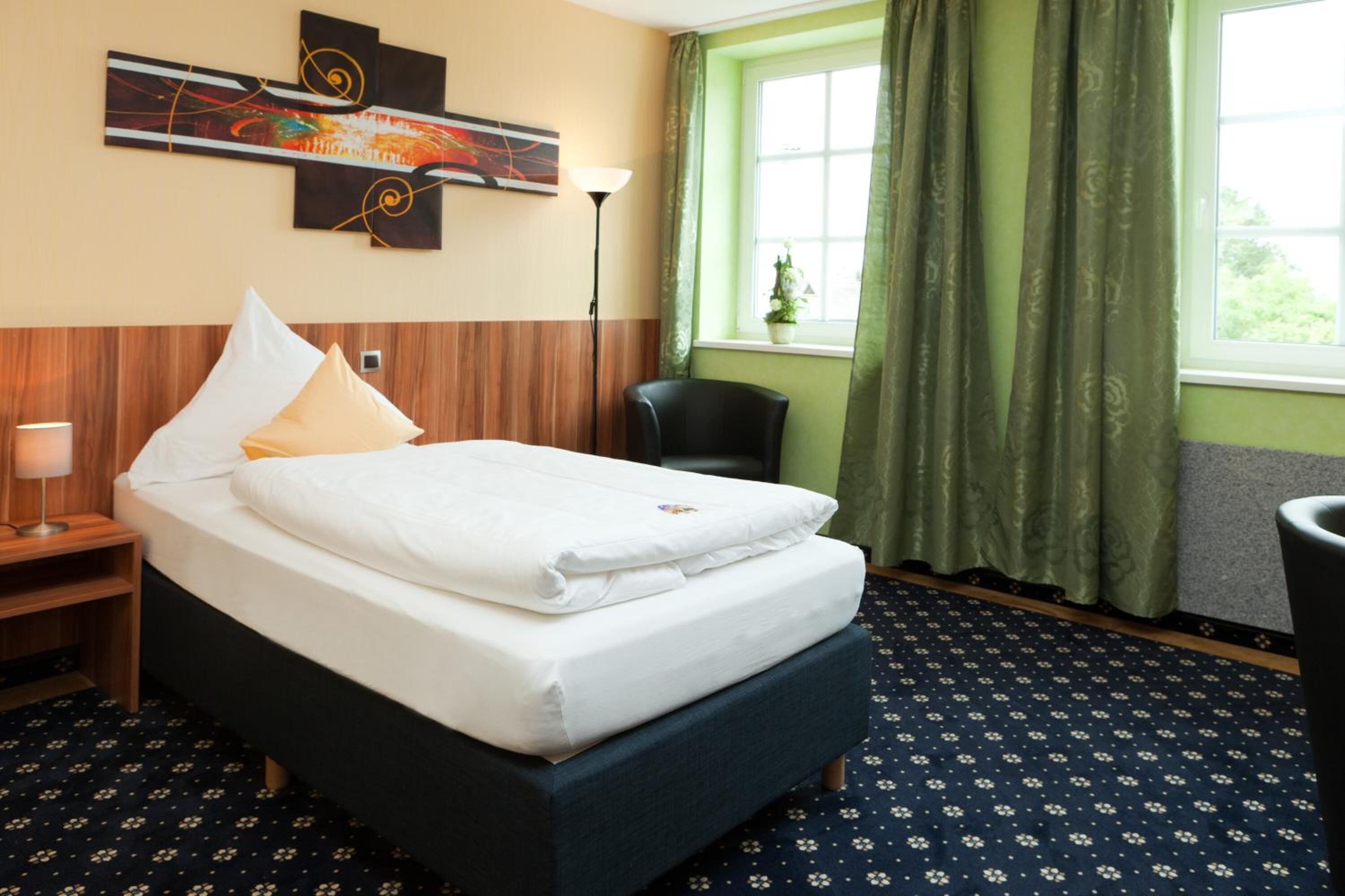 A hotel room with a bed, a chair, a lamp, a painting on the wall, and a window with curtains.