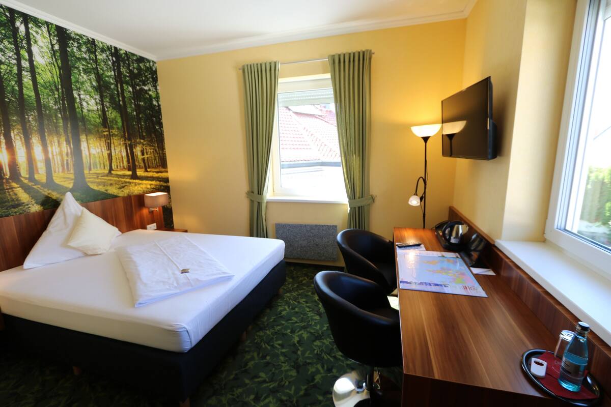 Habitación de hotel con cama, escritorio, silla y un ventanal con cortinas.