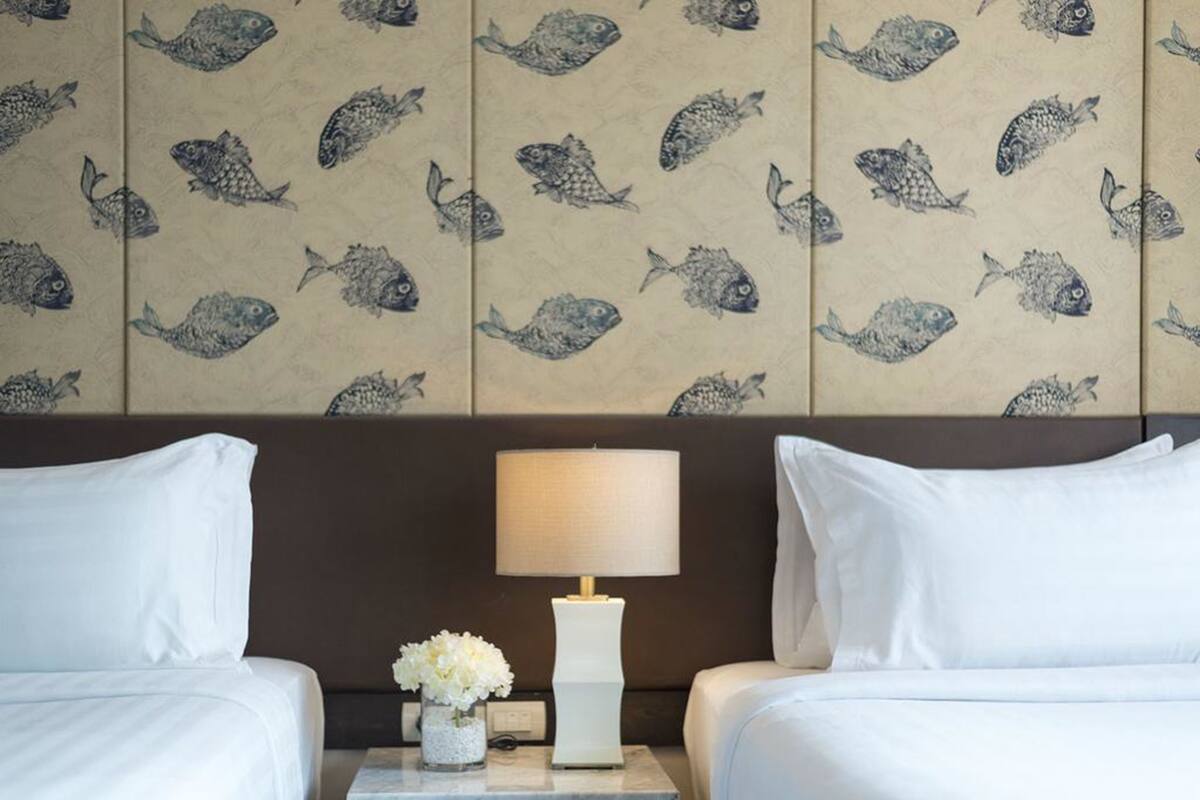 Quarto de hotel com duas camas, uma mesinha de cabeceira com abajur e uma parede com padrão de peixes.