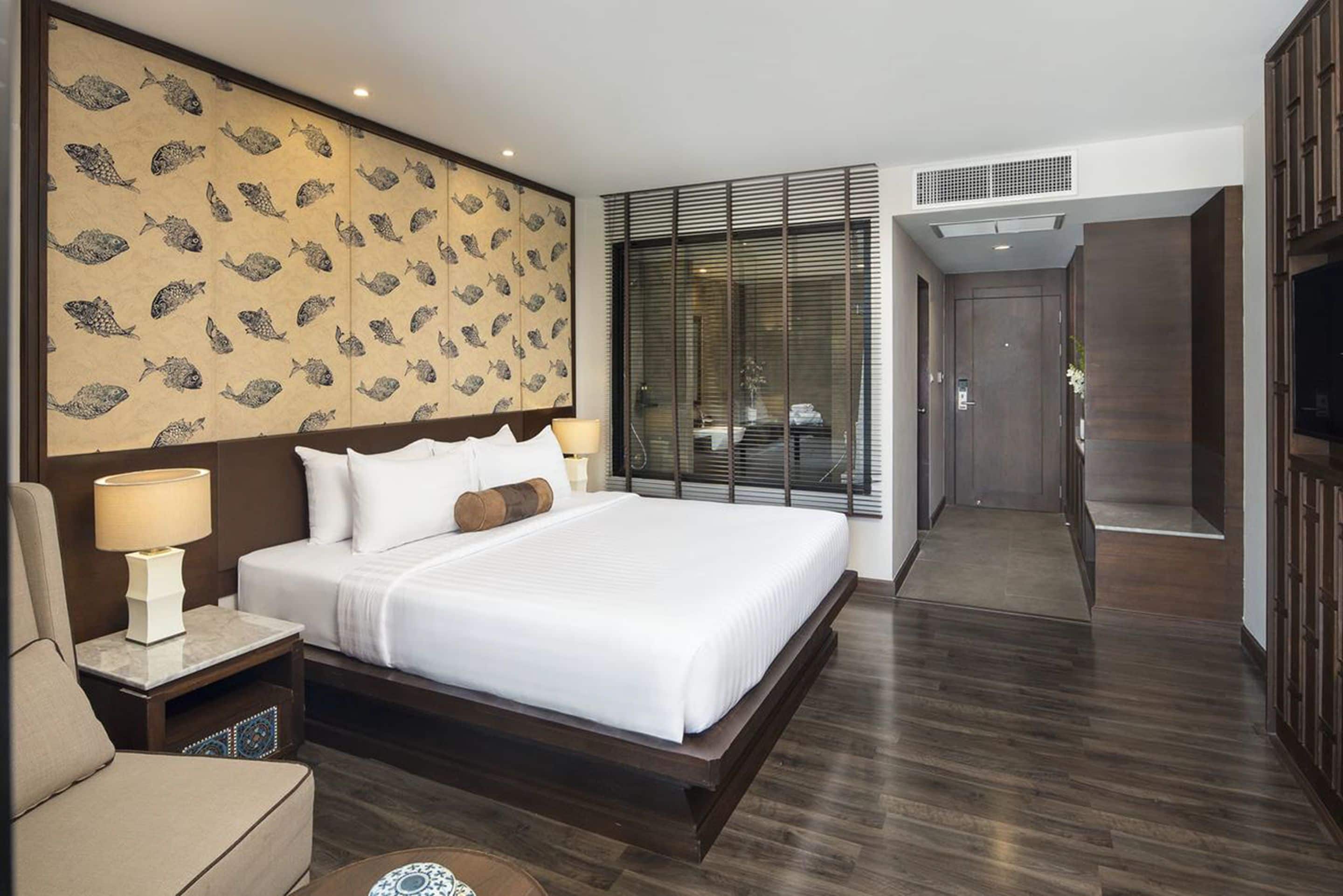 A modern hotel room with a large bed, a nightstand, a lamp, a wall with a fish pattern, a large window with blinds, and a door leading to another room.