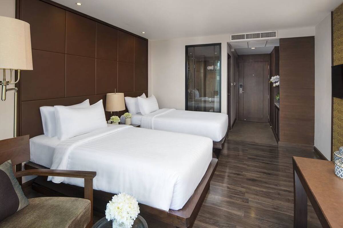 Quarto de hotel com duas camas, um espaço de estar com uma poltrona, uma mesinha e uma luminária.