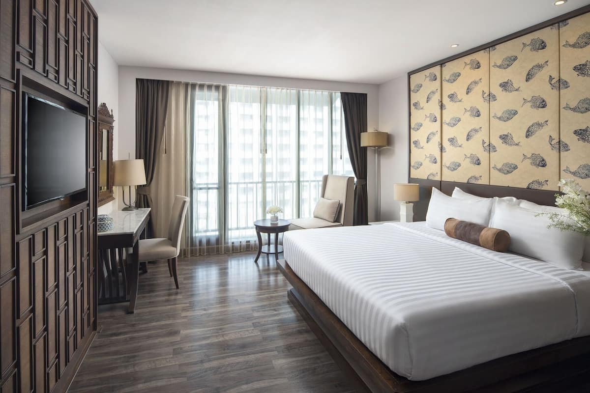 Quarto de hotel com uma cama grande, uma escrivaninha, duas cadeiras, uma televisão e vista para a cidade através de amplas janelas.