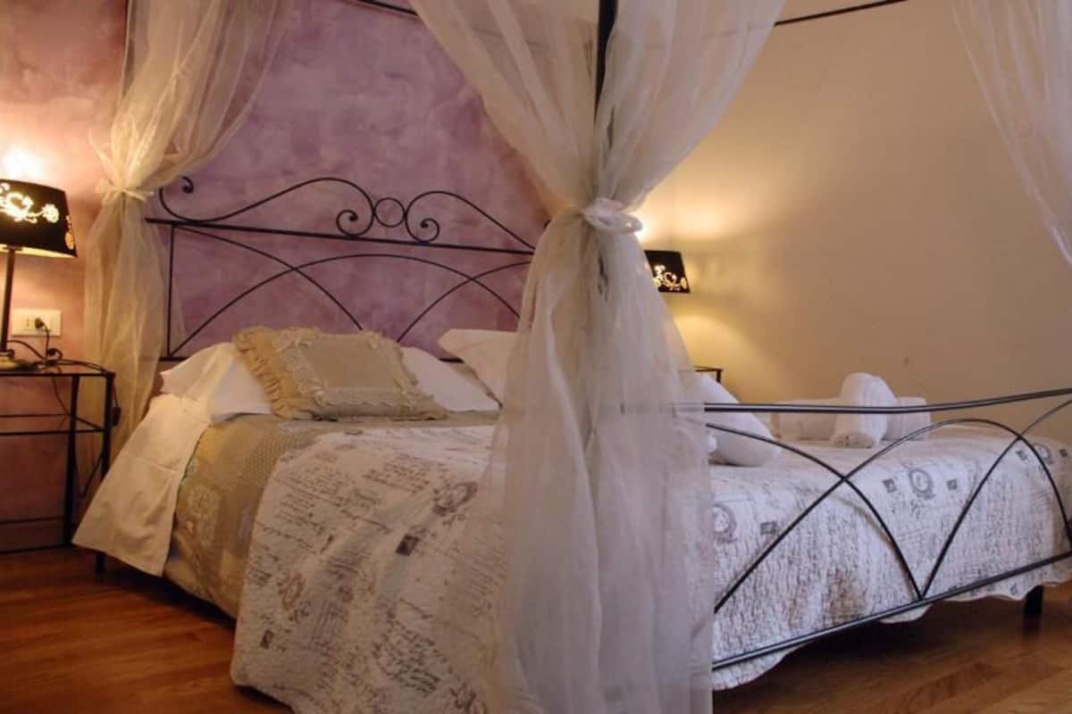A bedroom with a four-poster bed, a nightstand with a lamp, and a wall-mounted TV.