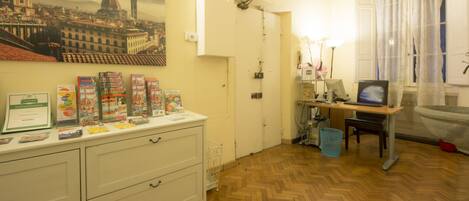 A room with a wooden herringbone floor, a desk with a computer, a chair, a small trash bin, and a wall-mounted shelf with brochures and certificates.