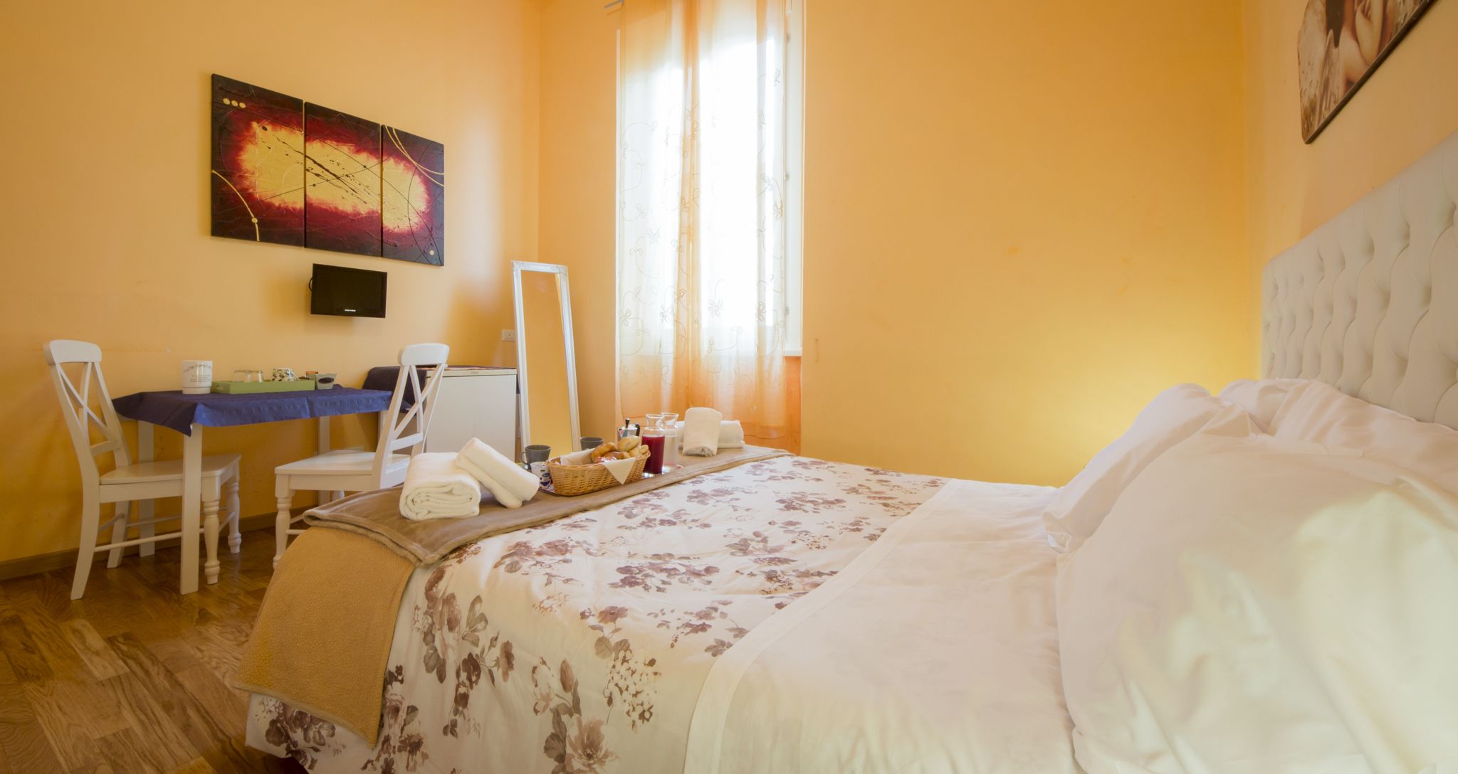 A hotel room with a bed, a desk with a chair, a mirror, and a painting on the wall.