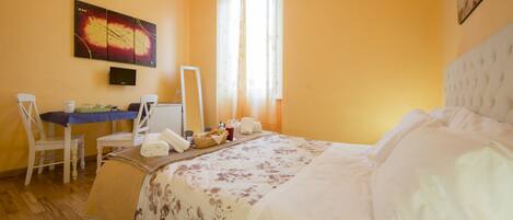 A hotel room with a bed, a desk with a chair, a mirror, and a painting on the wall.