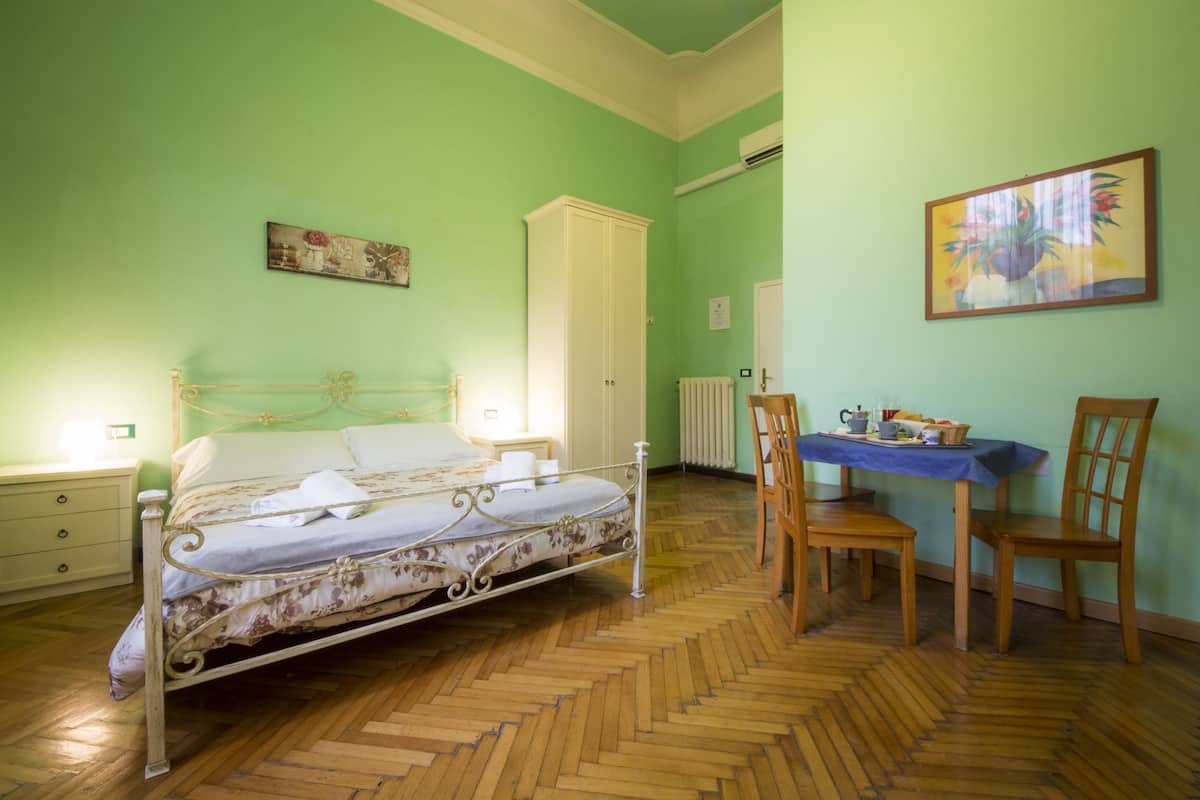 A bedroom with a bed, a wooden chair, a table with items, and a radiator.