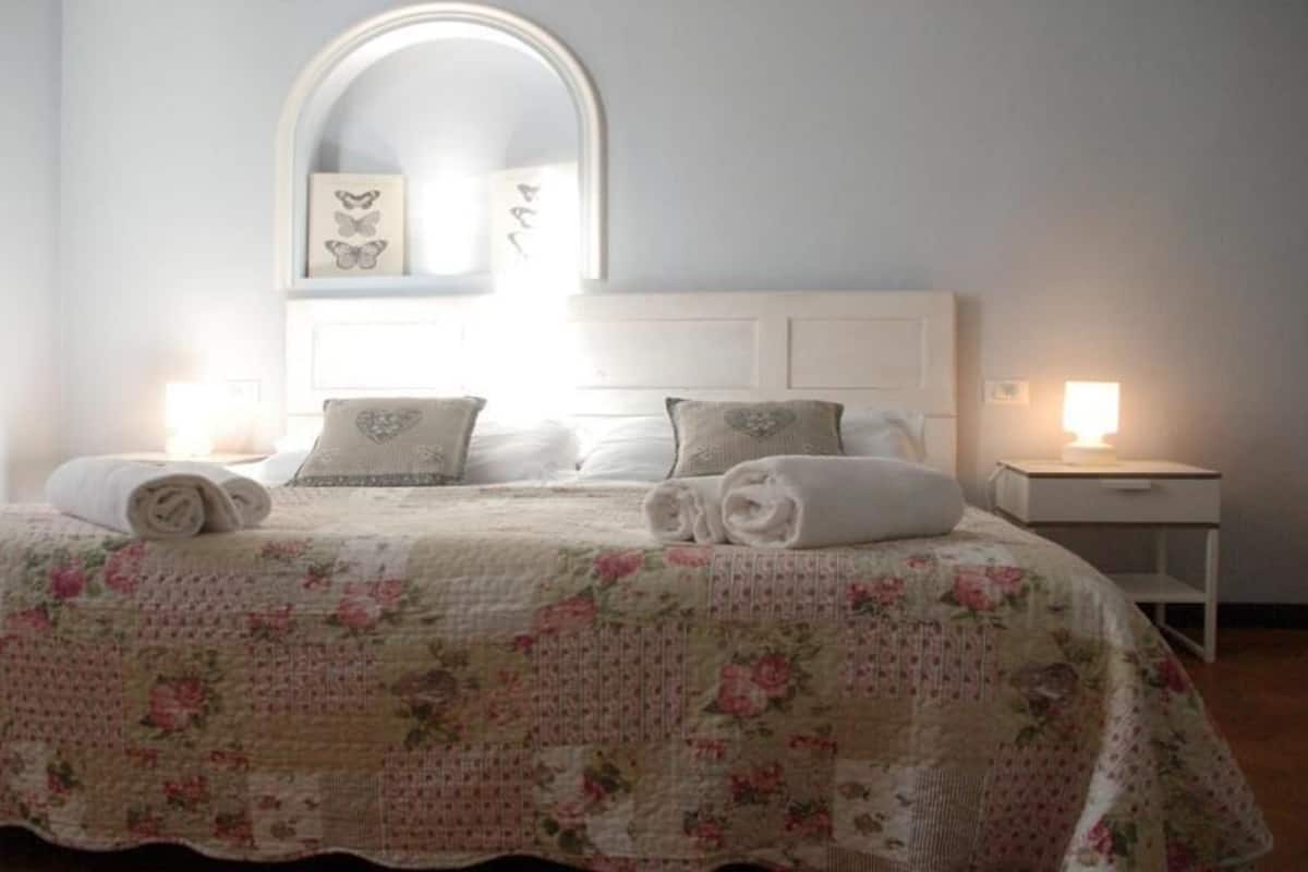 A neatly made bed with a floral bedspread, two pillows, and two rolled towels. There is a nightstand with a lamp on it.