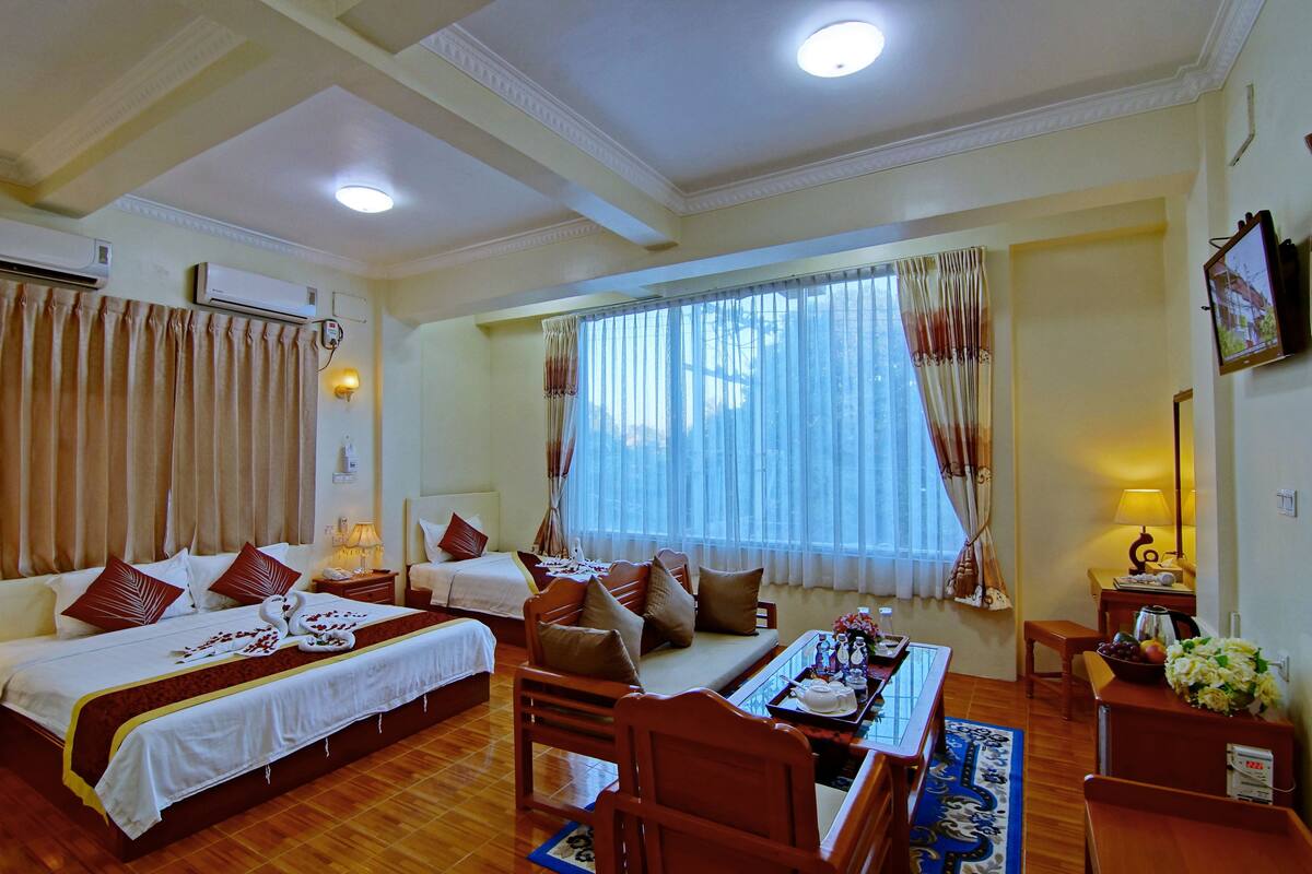 A hotel room with a large bed, a sofa, a dining table, and a window with curtains.