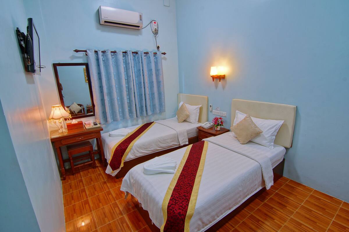 A hotel room with two beds, a desk with a lamp, a mirror, and an air conditioning unit.