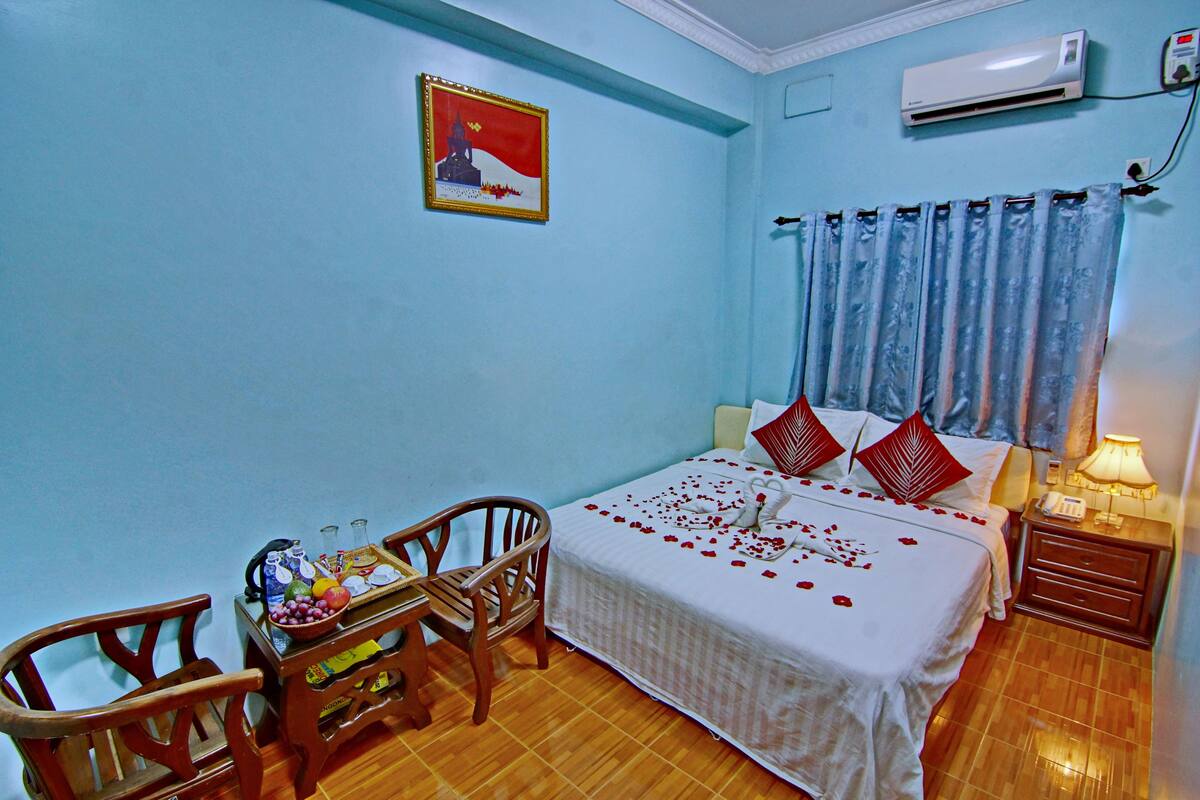 A bedroom with a bed, a nightstand, a chair, a table with a bowl of fruit, and a wall-mounted air conditioner.