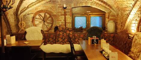 A dining area with a long wooden table, benches, a chandelier, and a stone wall with a window.