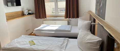 A hotel room with two beds, a flat-screen TV mounted on the wall, a window with brown curtains, and a radiator below the window.