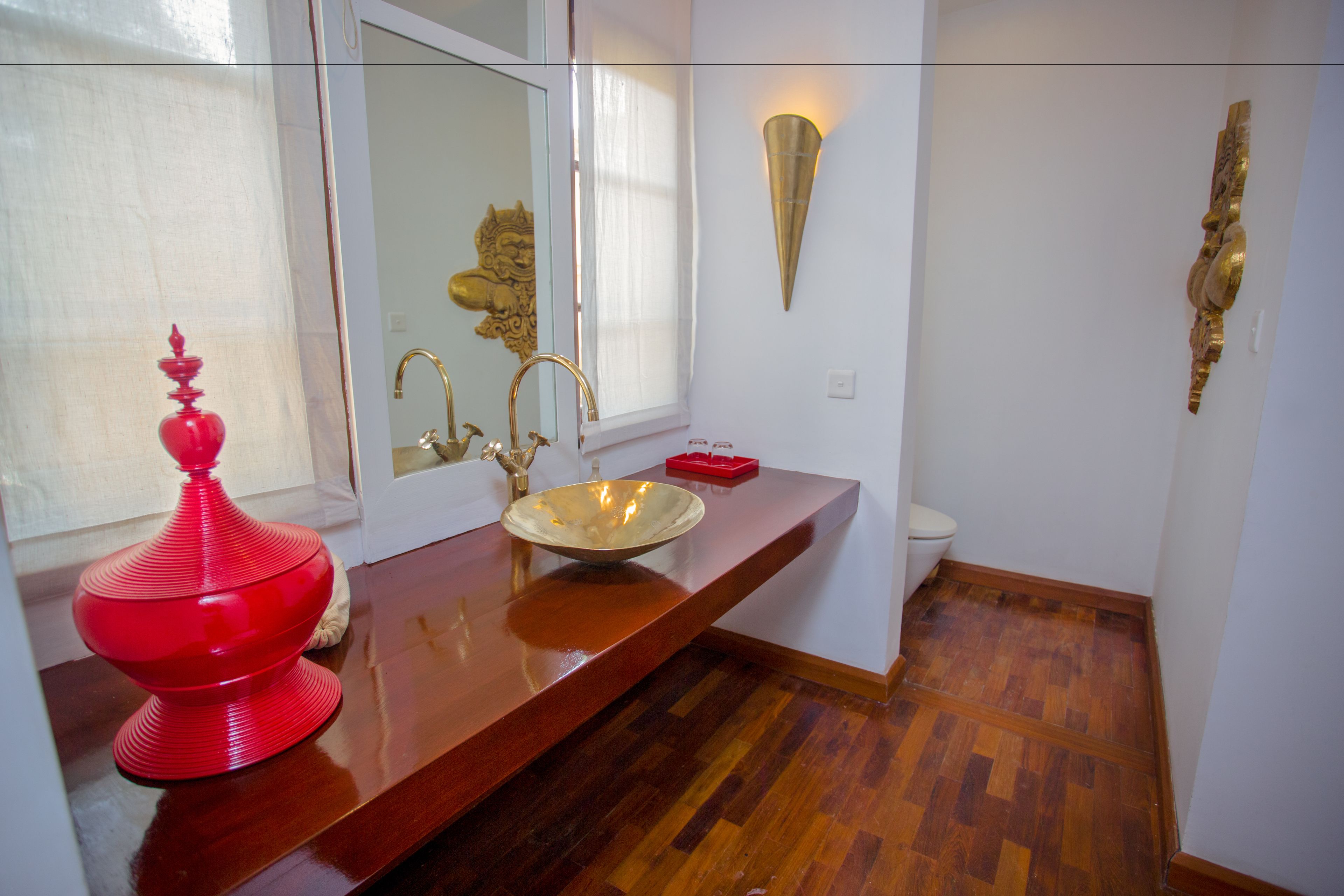 A bathroom with a wooden vanity, a red decorative item, a gold faucet, and a wall-mounted light fixture.