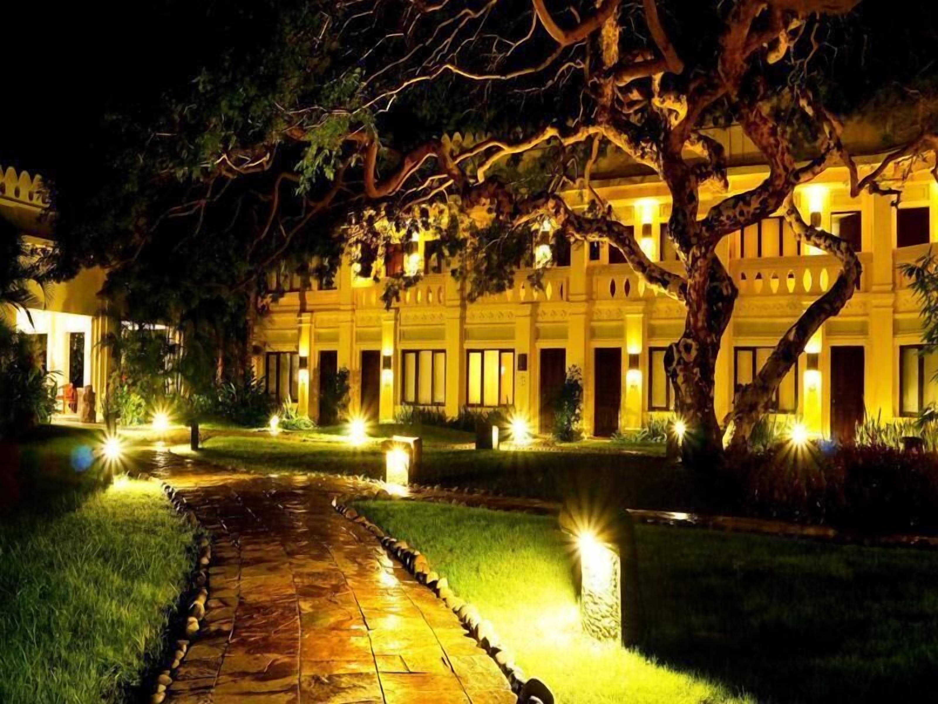 Ein gut beleuchtetes Hotel mit einem gepflasterten Weg zum Eingang, umgeben von Bäumen und beleuchtet durch Bodenleuchten.
