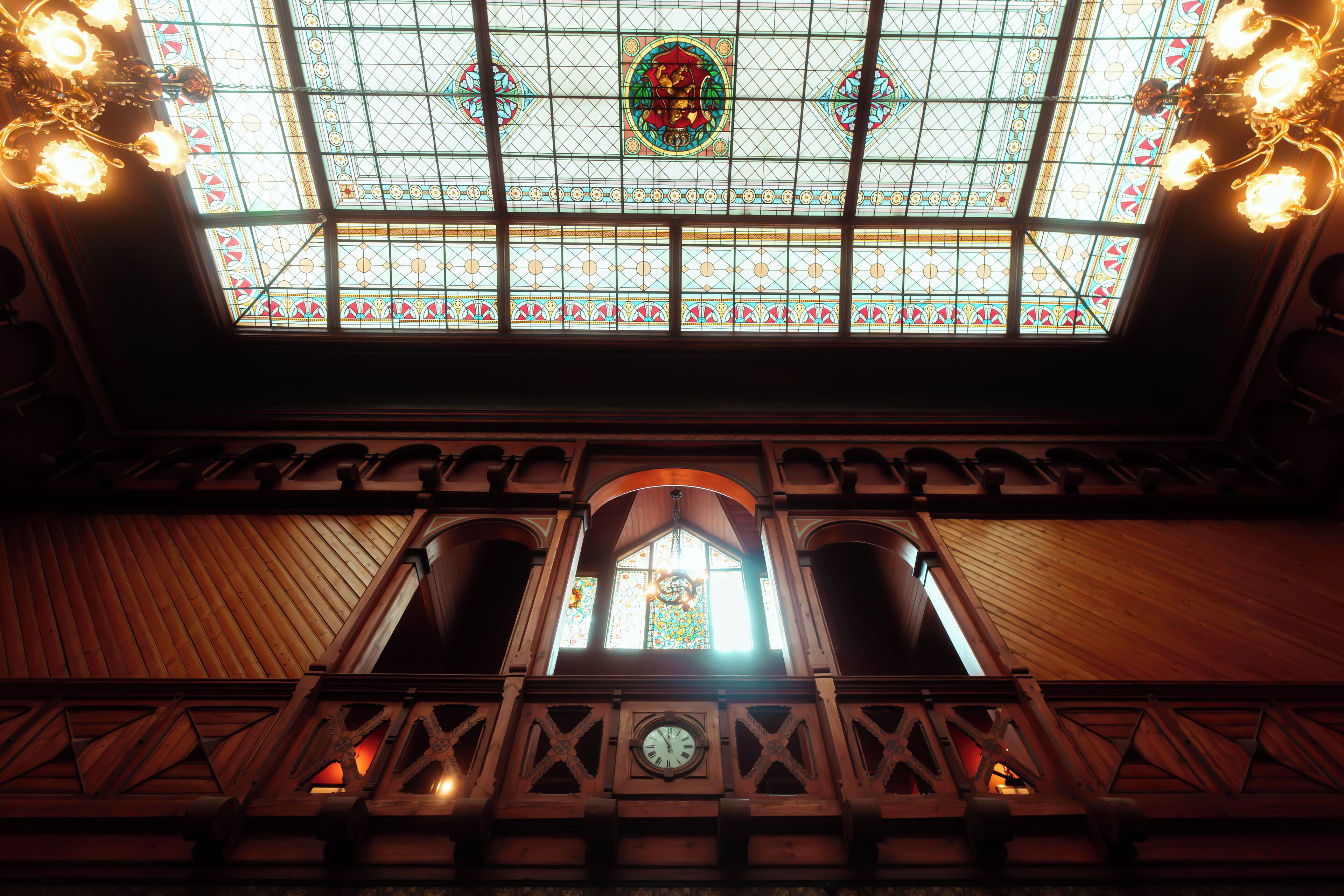 A stained glass window with geometric patterns and a central emblem, wooden beams, and a clock.