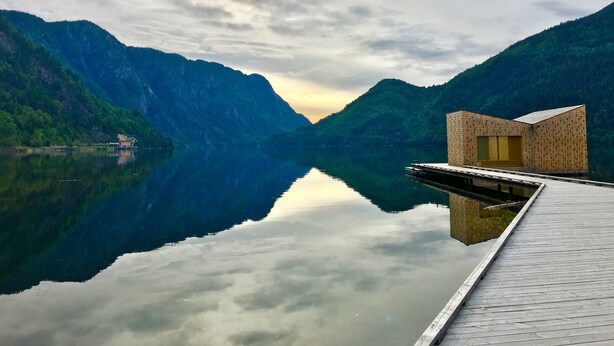 A serene lake with a wooden dock and a small brick building, surrounded by lush green mountains. - Dalen Hotel (Tokke)