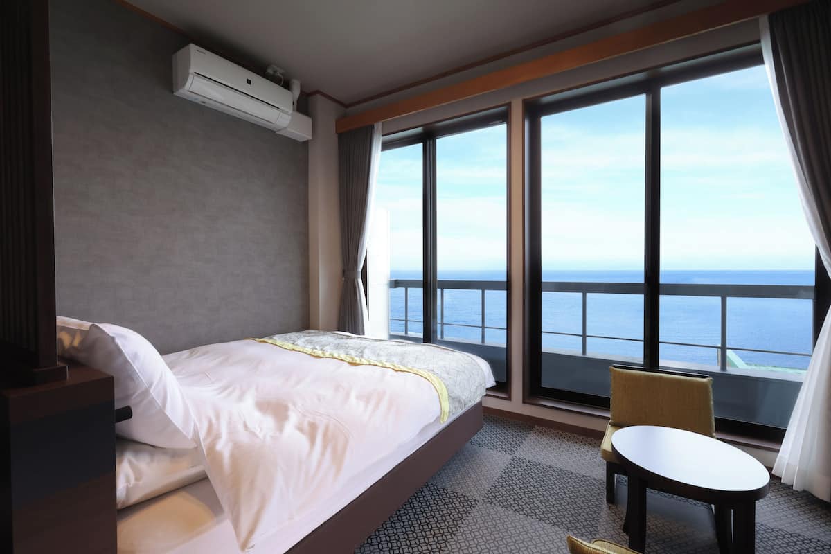 A hotel room with a bed, a small table, a chair, a large window with a view of the sea, and an air conditioning unit.