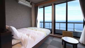 A hotel room with a bed, a small table, a chair, a large window with a view of the sea, and an air conditioning unit.