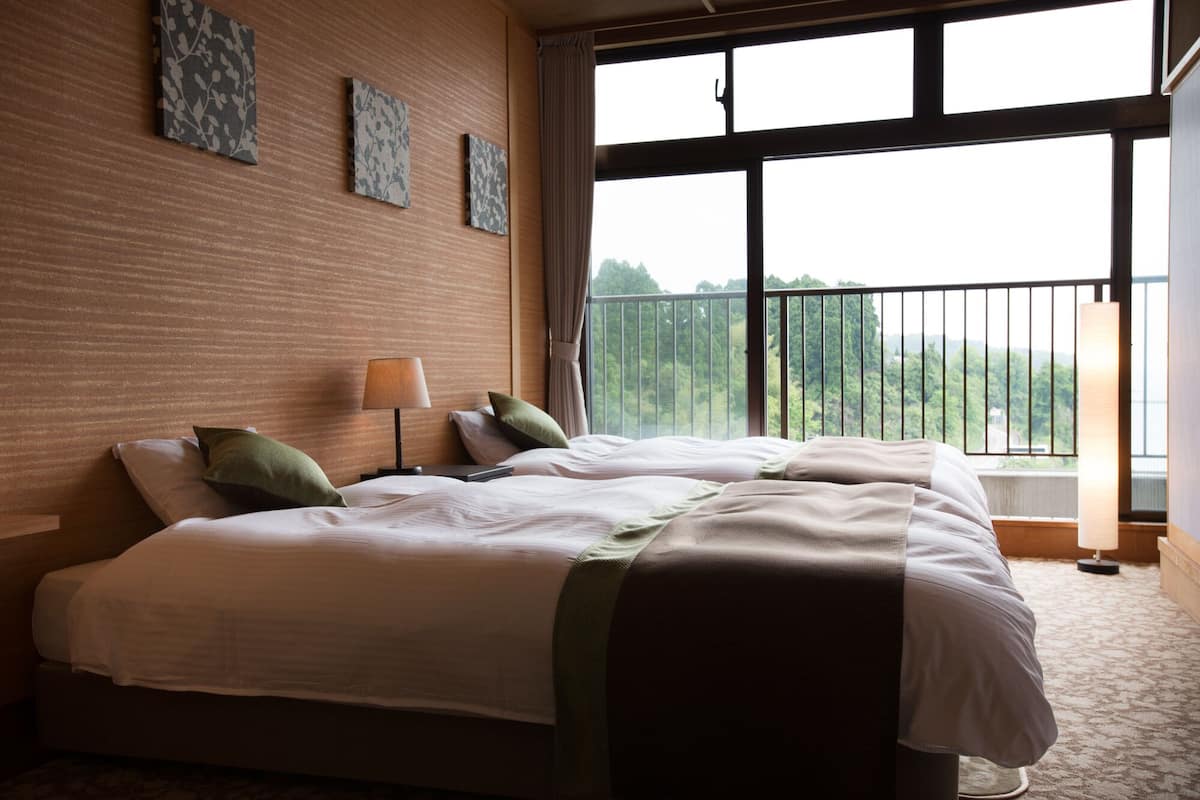 A hotel room with a large window, a bed with white bedding, two bedside tables, and a lamp.