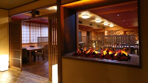 Una habitación japonesa tradicional con suelo de tatami, una mesa baja con cojines y vistas a un área de restaurante con una chimenea.