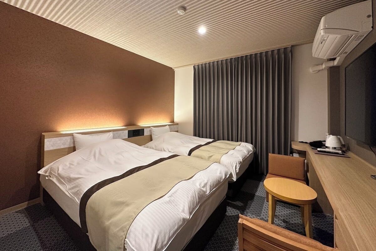 A hotel room with two beds, a desk, a chair, and a large window with curtains.