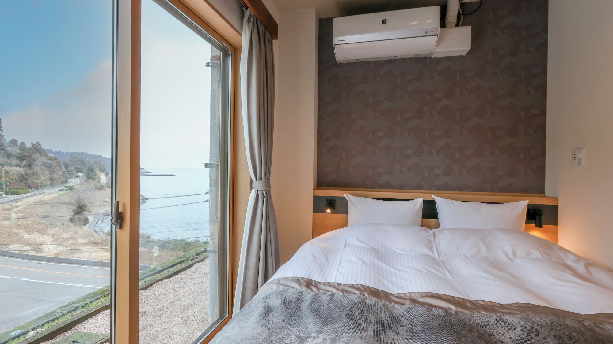 Habitación de hotel con un ventanal grande, cama con ropa de cama blanca, cabecera con interruptor de luz y unidad de aire acondicionado en la pared.