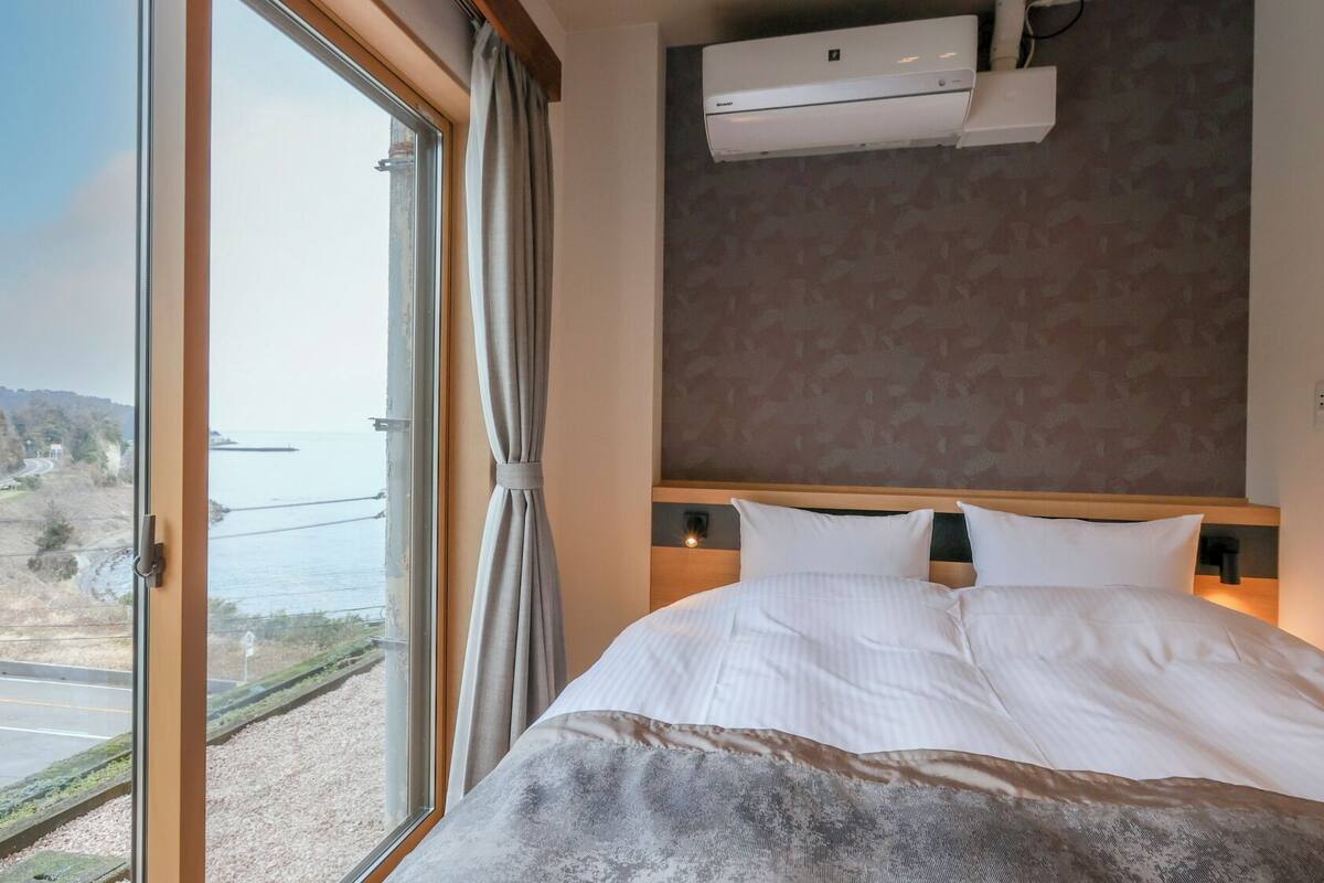 A hotel room with a large window, a bed with white bedding, a headboard with a light switch, and an air conditioning unit mounted on the wall.