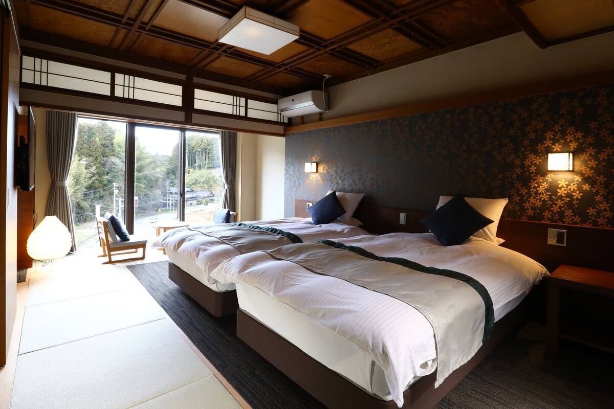A large bed with a wooden headboard, a bedside table, and a nightstand. There is a window with curtains, a chair, and a lamp.