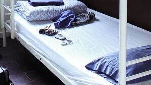 Una cama individual con estructura blanca, un cojín estampado con motivos florales, una manta azul y un par de gafas de sol sobre la cama.
