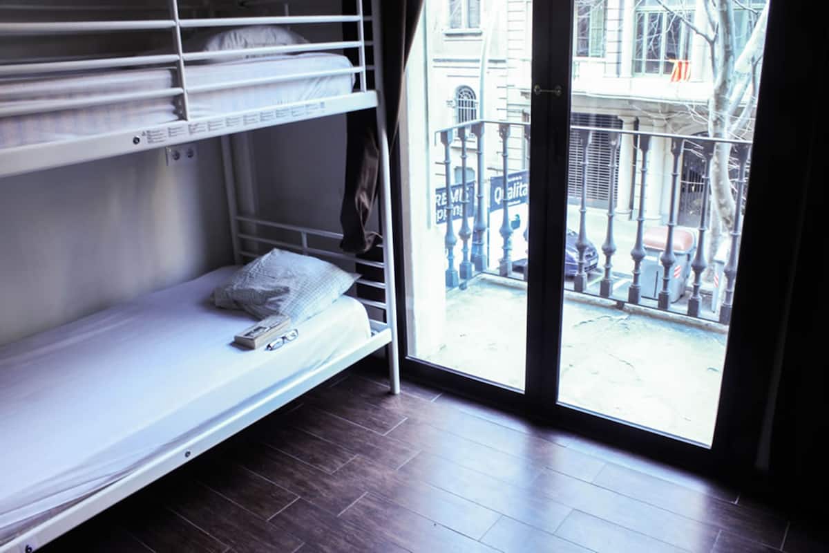 A dormitory-style room with bunk beds, a window with a view of a building and a balcony outside.