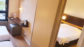 A compact hotel room with a bed, a small desk with a kettle, a TV, and a window with a view of the outside.