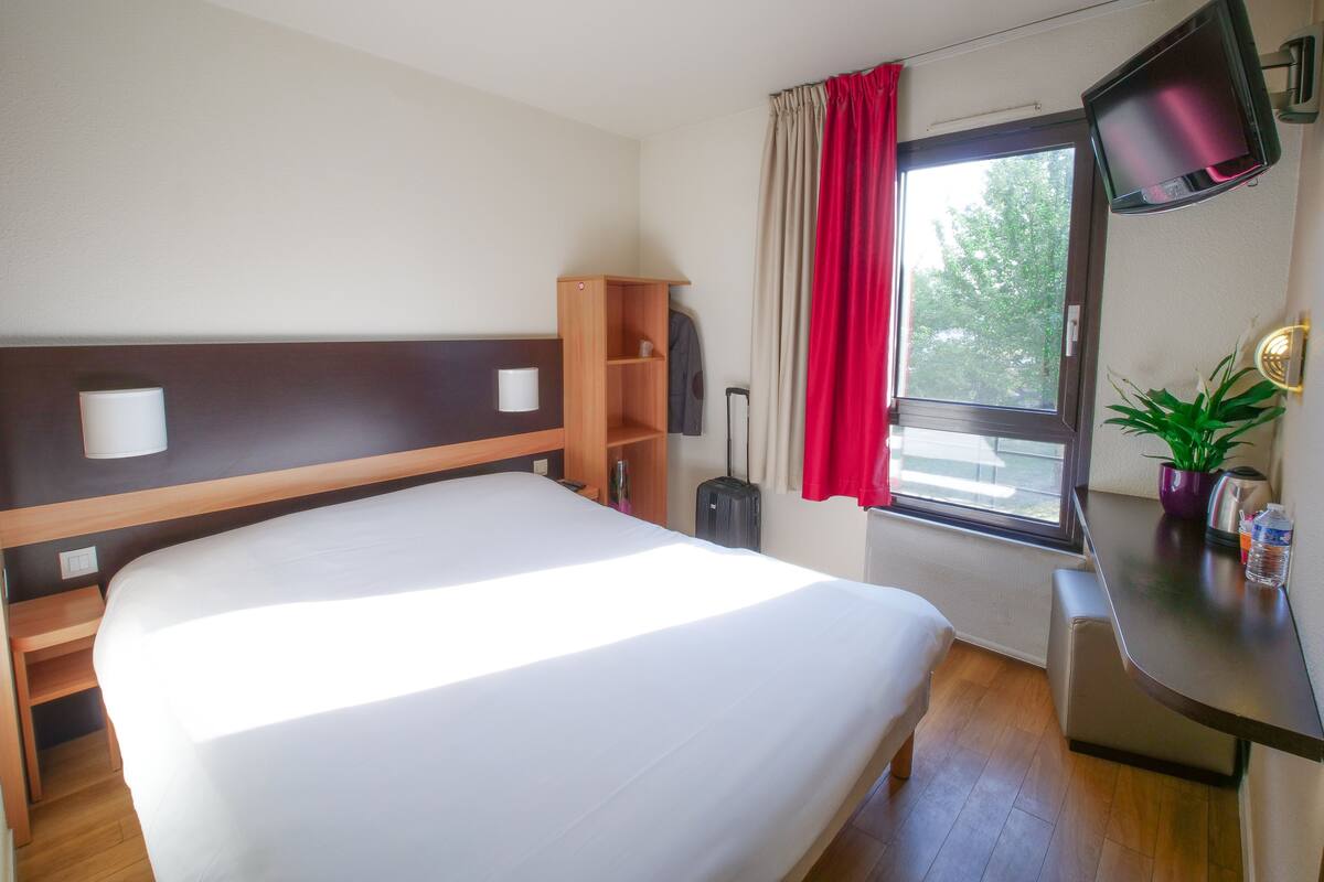 A hotel room with a bed, a wooden headboard, a desk with a TV, a window with curtains, and a potted plant.