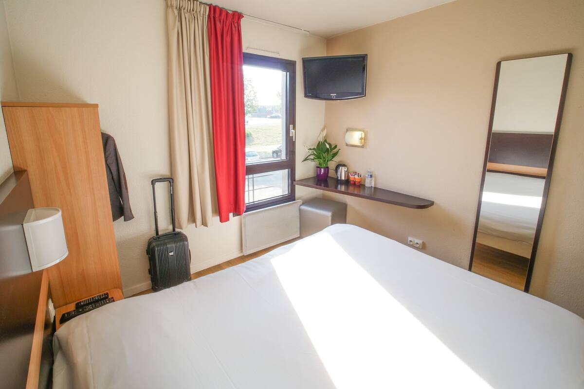 A hotel room with a bed, a desk, a TV, a mirror, a window with curtains, a plant, and a suitcase.